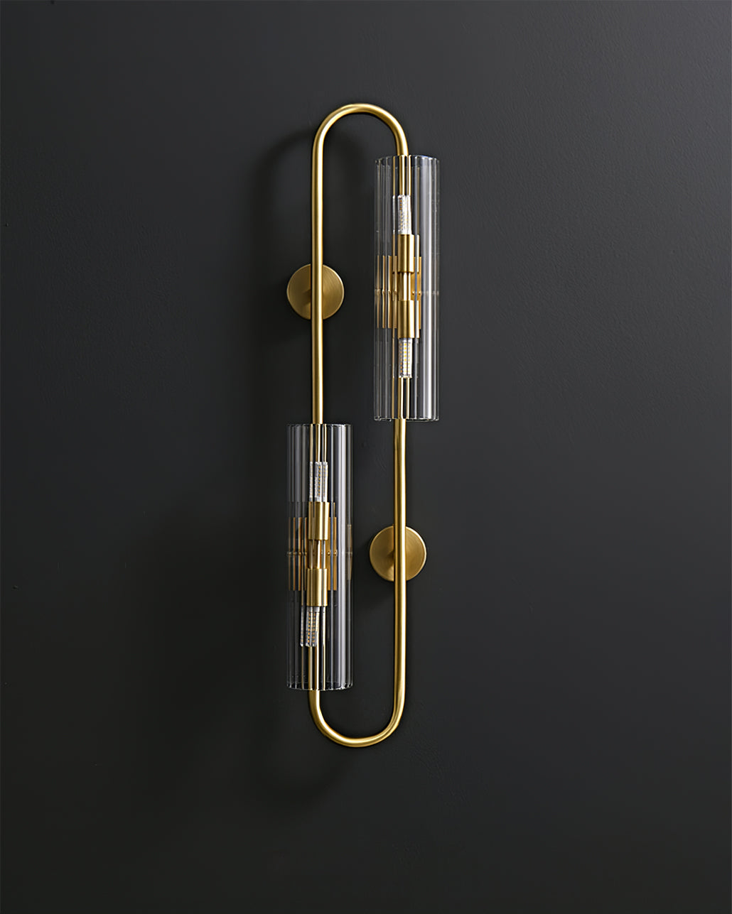 Unlit, this Clérachic brass wall light shows its two ribbed clear glass tubes and round wall plates against a dark background.