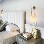 This Clérachic sconce lit beside a padded white headboard, with a grey-blue bedside table and a gold picture frame below.