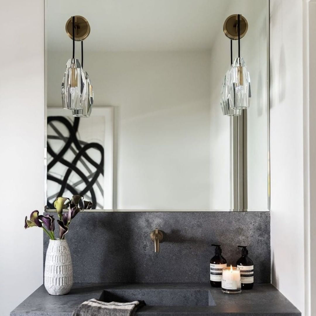 A pair of Clérachic crystal wall lights flanking a tall mirror above a dark stone vanity, with a vase of dark flowers and a lit candle.