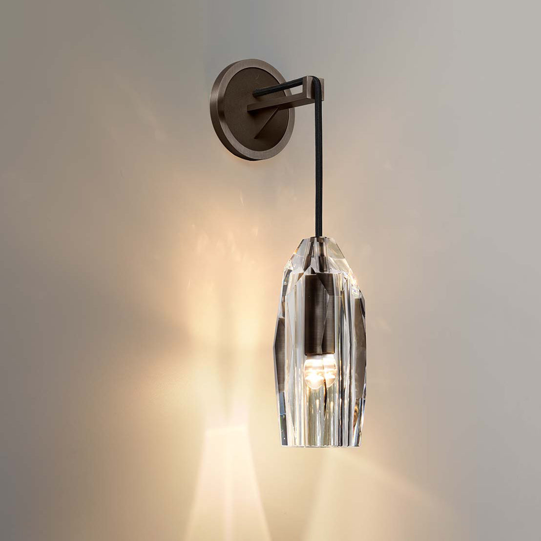 A lit wall light with a dark bronze-toned backplate and crystal shade, glowing against a warm grey wall, shown for this Clérachic range.