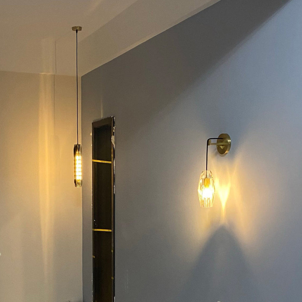 A Clérachic crystal wall light glowing amber against a blue-grey wall in a dim hallway, with a slim tube pendant light further back.