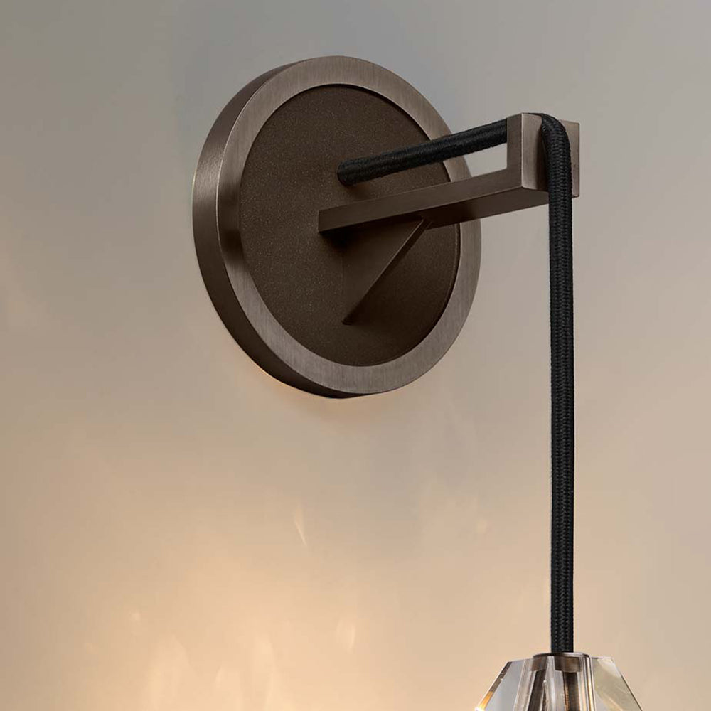 Close view of a dark bronze-toned round backplate and braided black cord on this Clérachic wall light, with the crystal top edge below.