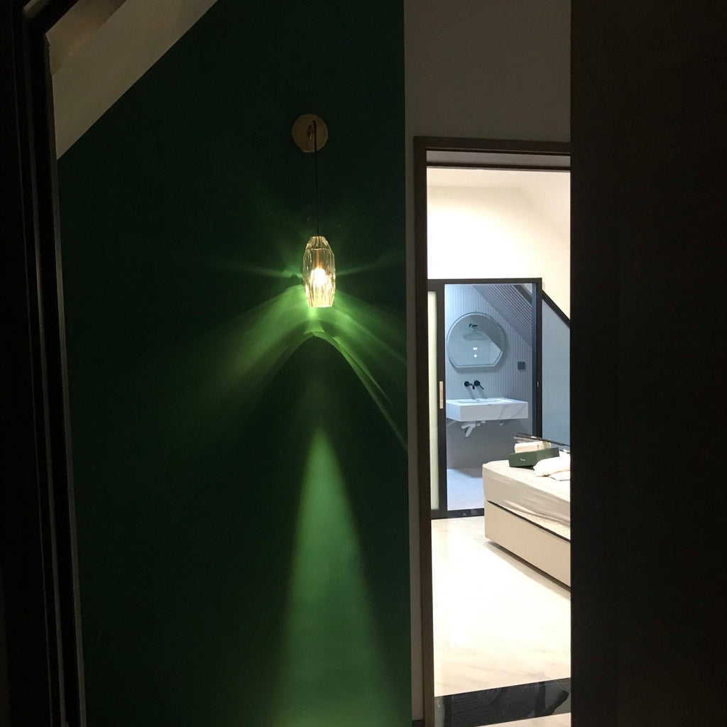 A Clérachic crystal wall light lit on a deep green wall, casting a green glow and a long beam down toward a bright bathroom doorway.