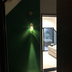 A Clérachic crystal wall light lit on a deep green wall, casting a green glow and a long beam down toward a bright bathroom doorway.