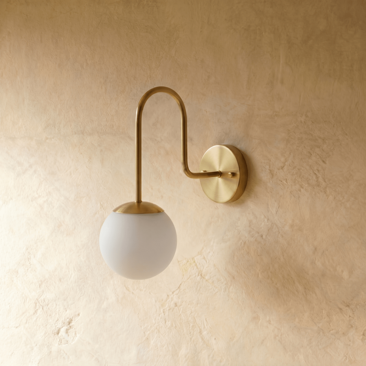 Clérachic wall lamp in brushed brass, the frosted glass globe glowing beneath a curved metal arm.