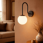 Clérachic wall lamp in matte black lighting a living room corner beside a sofa and dried-grass vase.