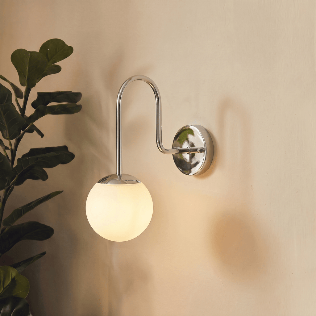 Clérachic wall lamp in chrome beside a leafy fiddle-leaf fig plant against a plaster wall.