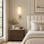 A Clérachic frosted glass wall sconce lit above a dark wood nightstand with brass handles, next to a channelled cream headboard.