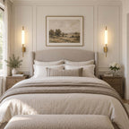 Two Clérachic brass wall sconces lit either side of a landscape painting above a neatly made bed with a linen quilt.