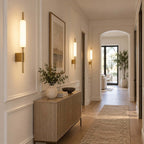 A row of Clérachic brass wall sconces lit along a panelled hallway, with a wooden sideboard and an arched doorway at the far end.