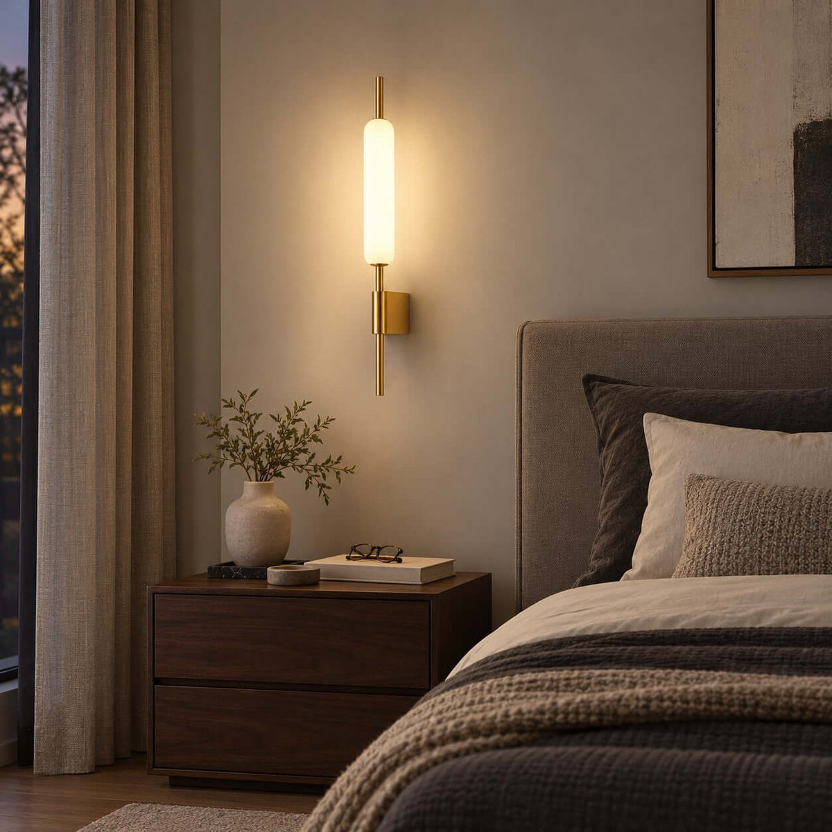 Lit above a dark nightstand and a vase of pale branches, this Clérachic brass wall sconce sits near a grey headboard and heavy curtains.