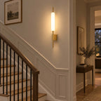 This Clérachic brass wall sconce lights a staircase landing, beside a wooden banister and a small console table with a plant.