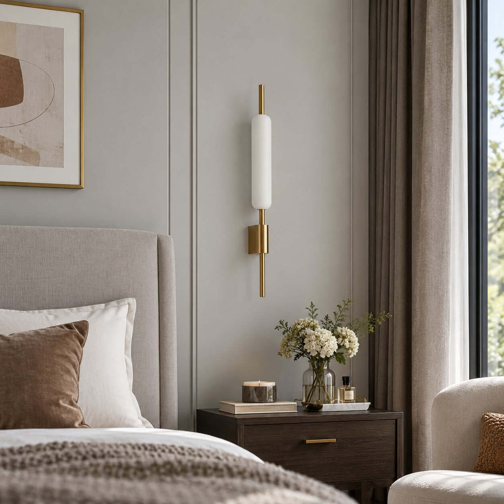 Unlit, this Clérachic wall sconce shows its white glass tube and brass back plate beside a grey bed and a vase of cream flowers.