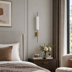 Unlit, this Clérachic wall sconce shows its white glass tube and brass back plate beside a grey bed and a vase of cream flowers.