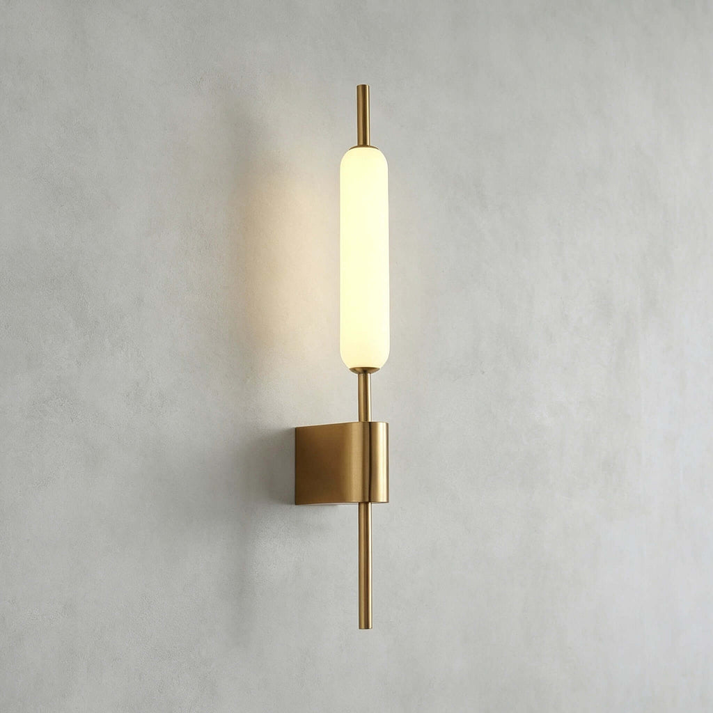 Straight-on view of this Clérachic brass wall sconce lit on a grey plaster wall, its glass tube above a square brass mount.
