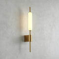 Straight-on view of this Clérachic brass wall sconce lit on a grey plaster wall, its glass tube above a square brass mount.