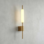 Straight-on view of this Clérachic brass wall sconce lit on a grey plaster wall, its glass tube above a square brass mount.
