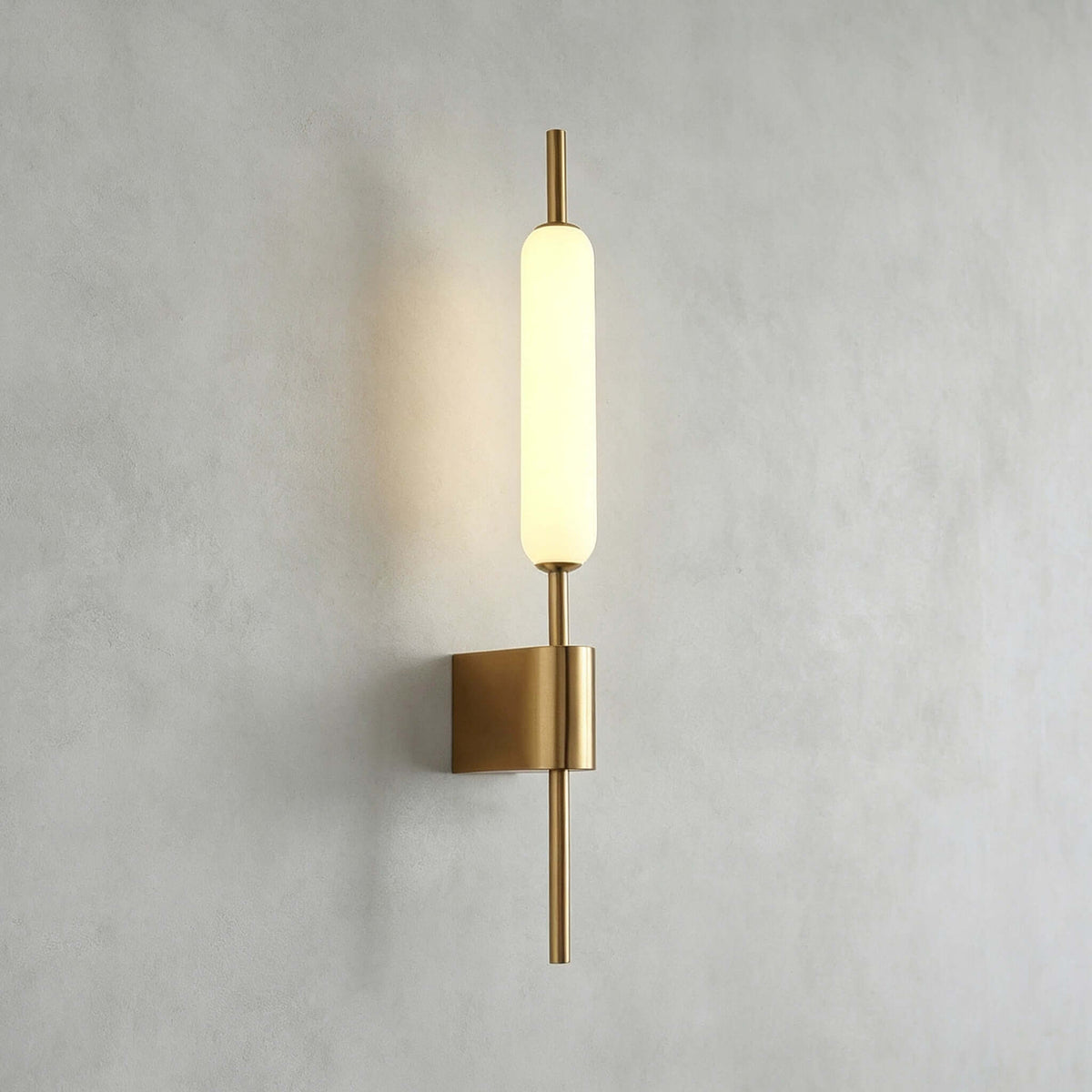 Straight-on view of this Clérachic brass wall sconce lit on a grey plaster wall, its glass tube above a square brass mount.