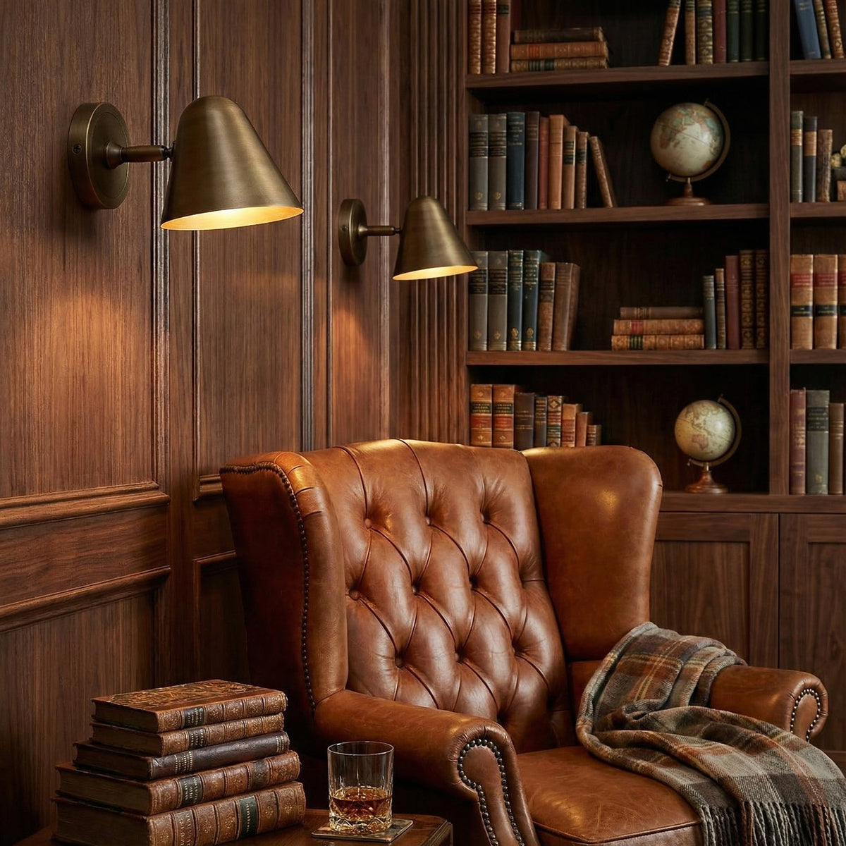 Two Clérachic antique brass wall lights on a wood-panelled wall above a tan leather armchair, with a tall bookcase and a whisky glass.