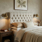 Two Clérachic antique brass wall lights either side of a tufted linen headboard, above wooden bedside tables, in a calm bedroom.