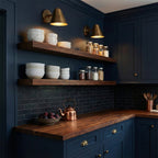 Two Clérachic antique brass wall lights above open wooden shelves of bowls and jars, against navy kitchen cabinets and a dark tiled wall.