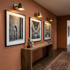 Three Clérachic antique brass wall lights in a row above framed black-and-white prints and a console table, on a terracotta wall.