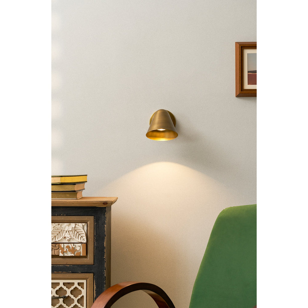 A Clérachic antique brass wall light on a pale wall above a painted cabinet, with a green chair and a small framed print nearby.
