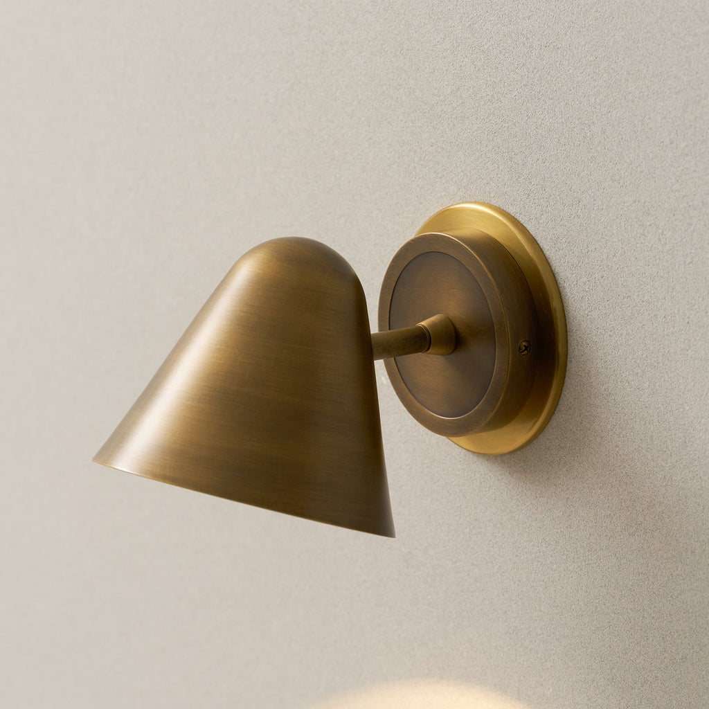 Close view of a Clérachic antique brass wall light on a cream wall, with the brass cone shade angled down from its round backplate.
