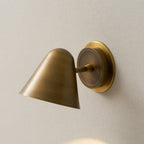 Close view of a Clérachic antique brass wall light on a cream wall, with the brass cone shade angled down from its round backplate.