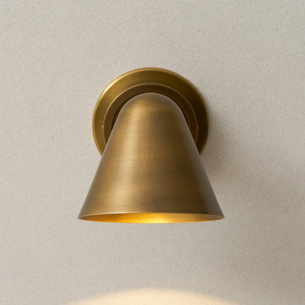 Front view of the cone shade of this Clérachic antique brass wall light, with warm light glowing at its lower rim.