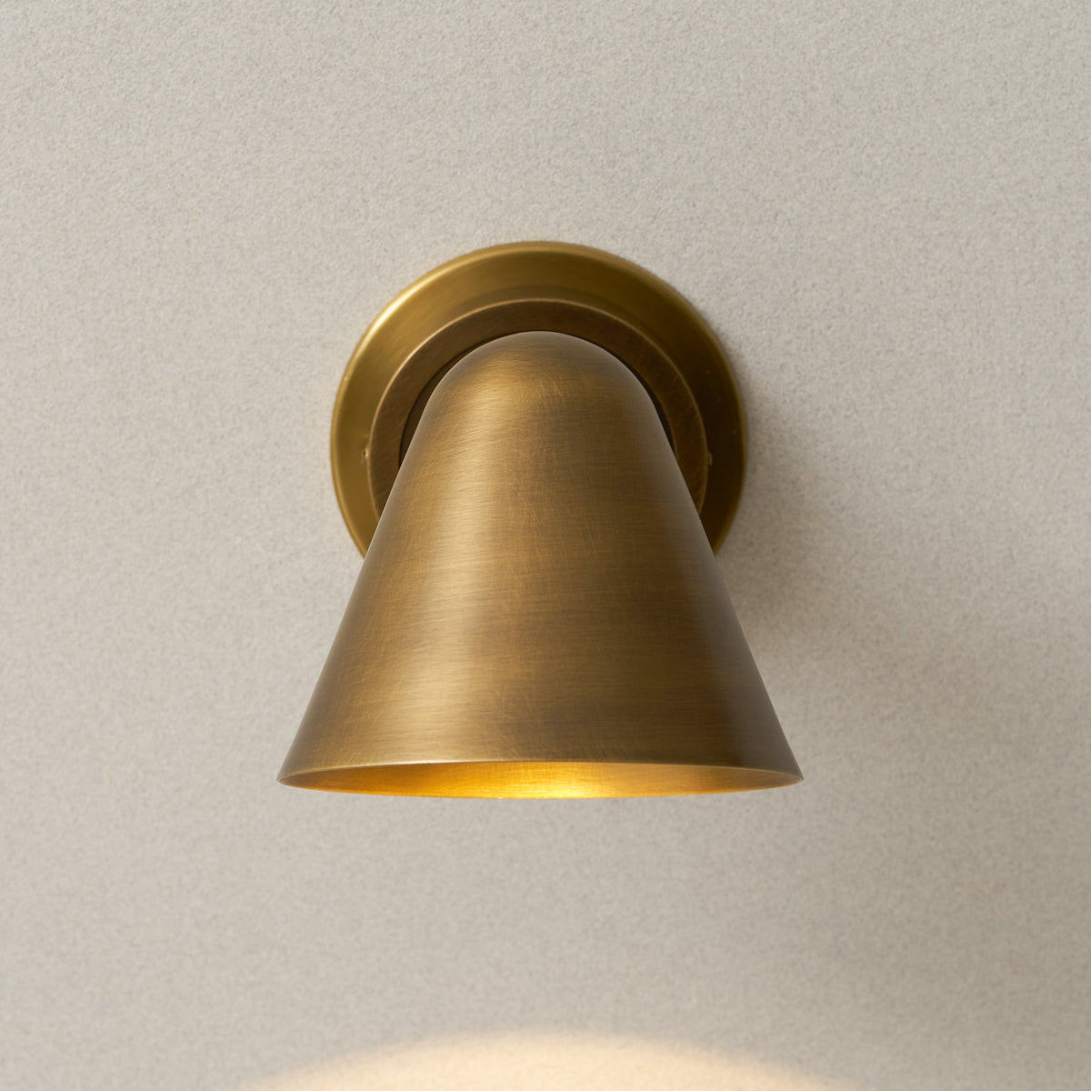 Front view of the cone shade of this Clérachic antique brass wall light, with warm light glowing at its lower rim.