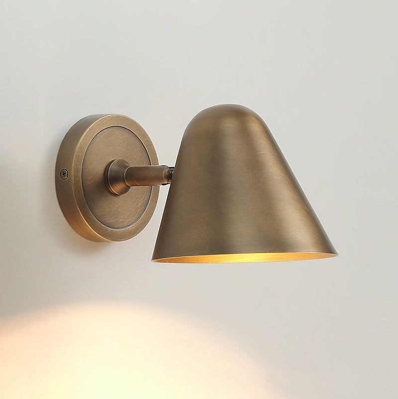 A Clérachic antique brass wall light on a pale wall, seen from the side with its short arm, round backplate and a pool of warm light below.