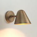 A Clérachic antique brass wall light on a pale wall, seen from the side with its short arm, round backplate and a pool of warm light below.