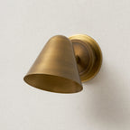 Close view of a Clérachic antique brass wall light angled down on a cream wall, showing the brushed finish on the cone shade.