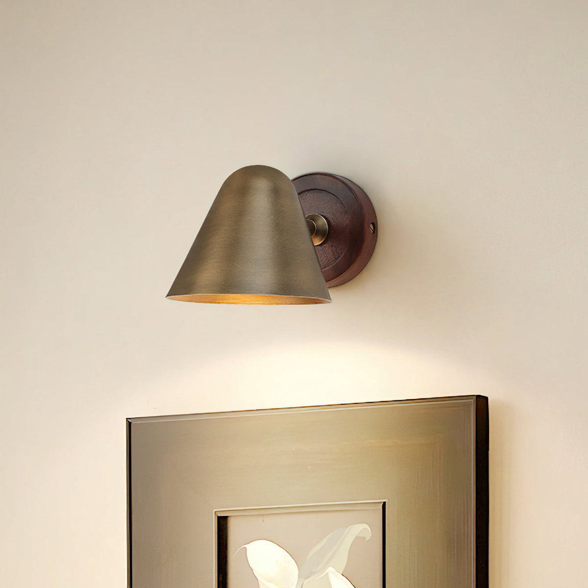 A Clérachic wall light with a brass cone shade and a dark wooden backplate on a cream wall above a framed picture, with a warm glow below.