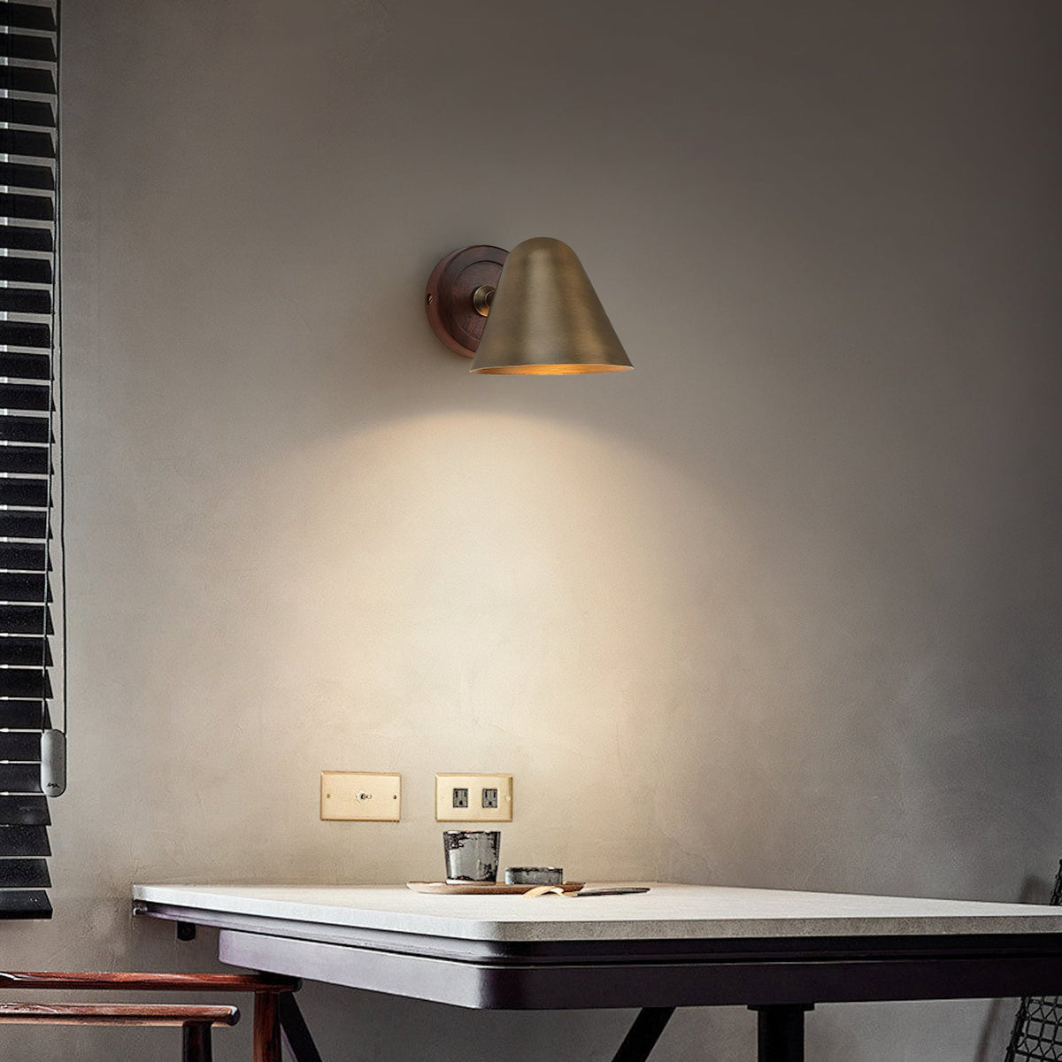 A Clérachic wall light with a brass cone shade and a dark wooden backplate on a grey wall above a small table and a slatted blind.