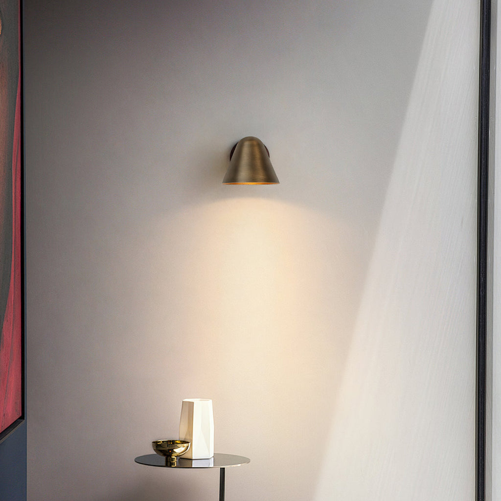 A small Clérachic brass wall light casting a soft cone of light on a grey wall above a slim side table with a vase and a bowl.