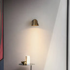 A small Clérachic brass wall light casting a soft cone of light on a grey wall above a slim side table with a vase and a bowl.