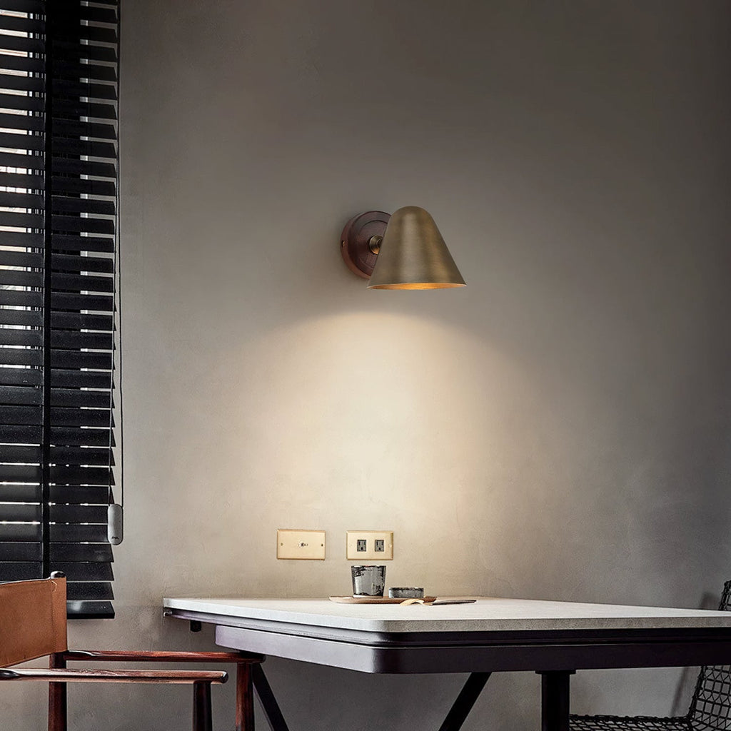 A Clérachic wall light with a brass shade and a dark wooden backplate on a grey wall above a white-topped table, beside a slatted blind.