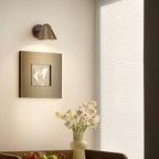 A Clérachic wall light with a brass shade on a cream wall above a framed print and a vase of white flowers, with a pale window blind.