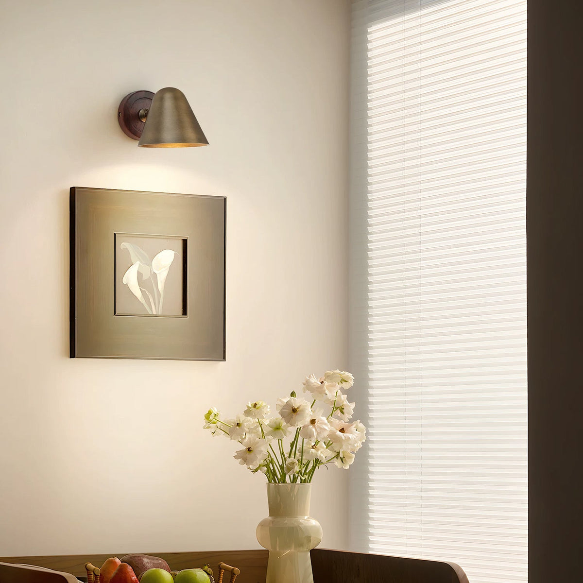 A Clérachic wall light with a brass shade on a cream wall above a framed print and a vase of white flowers, with a pale window blind.