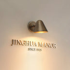 A Clérachic brass wall light lighting a wall with raised lettering below it, with a soft warm pool of light on the plaster.
