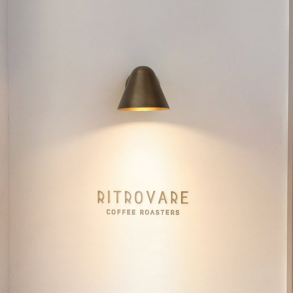 A Clérachic brass wall light high on a pale wall, casting a warm cone of light down over a small wall sign.