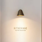A Clérachic brass wall light high on a pale wall, casting a warm cone of light down over a small wall sign.