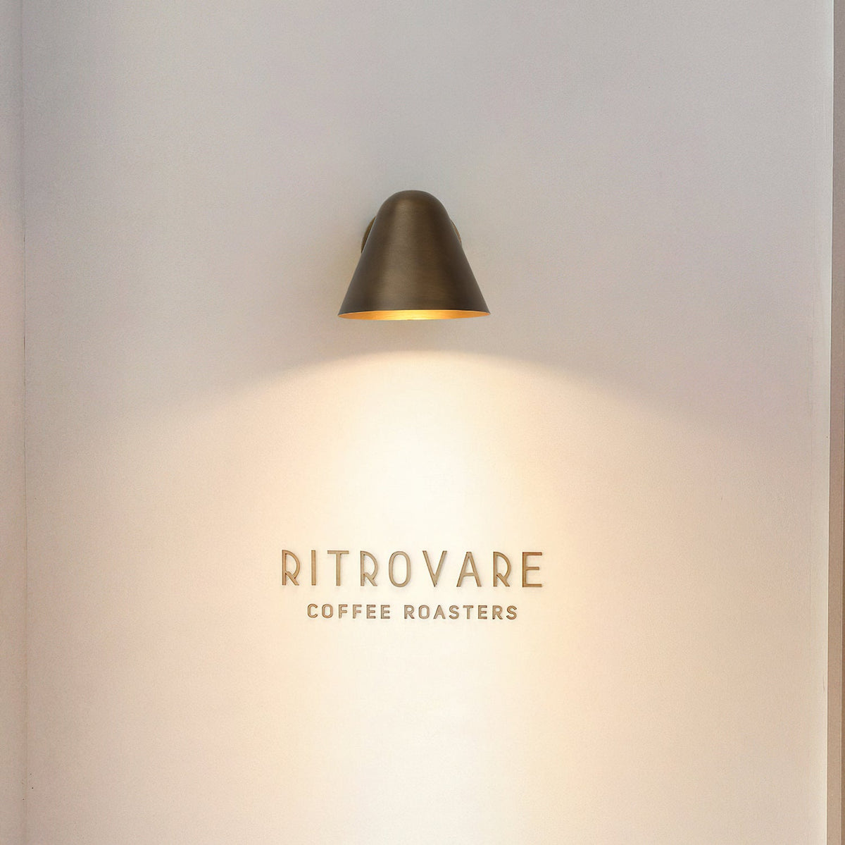 A Clérachic brass wall light high on a pale wall, casting a warm cone of light down over a small wall sign.