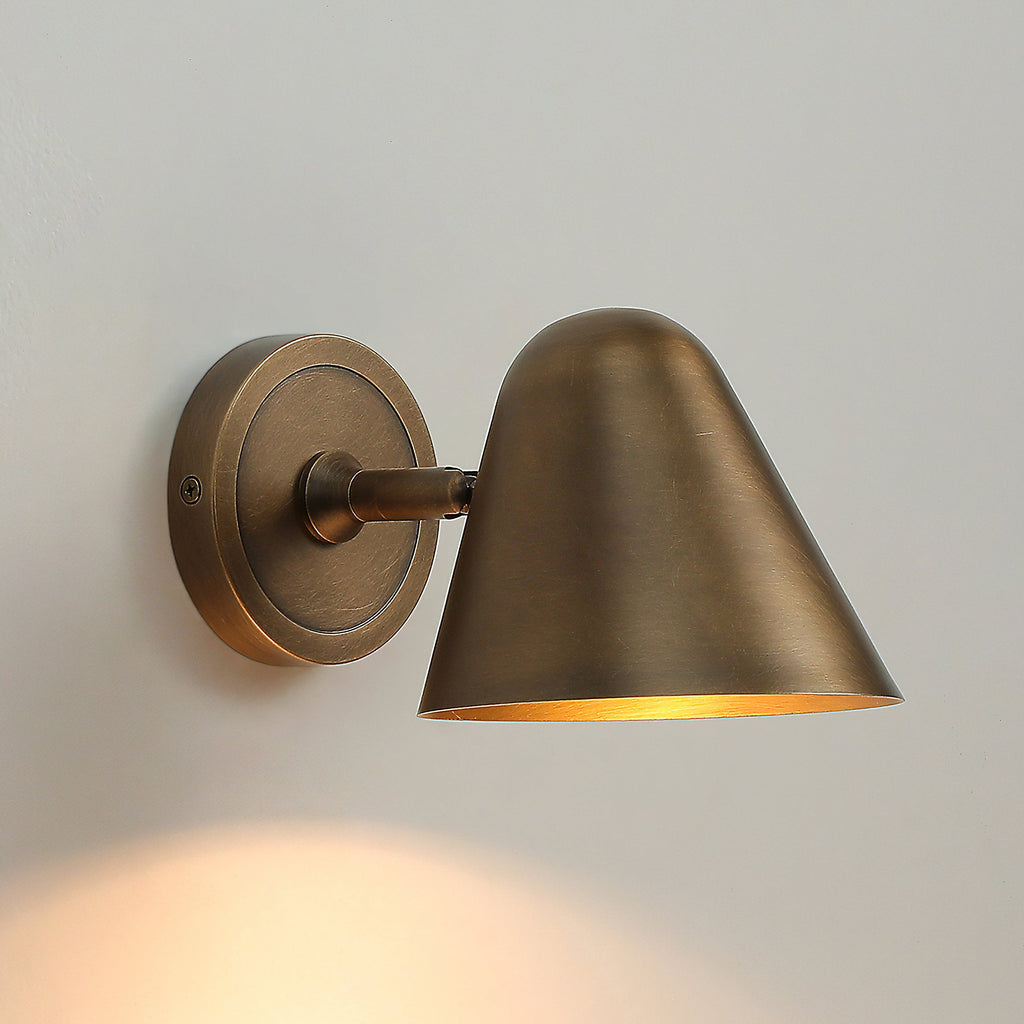 A Clérachic antique brass wall light with a round brass backplate, lit and casting warm light down a pale grey wall.