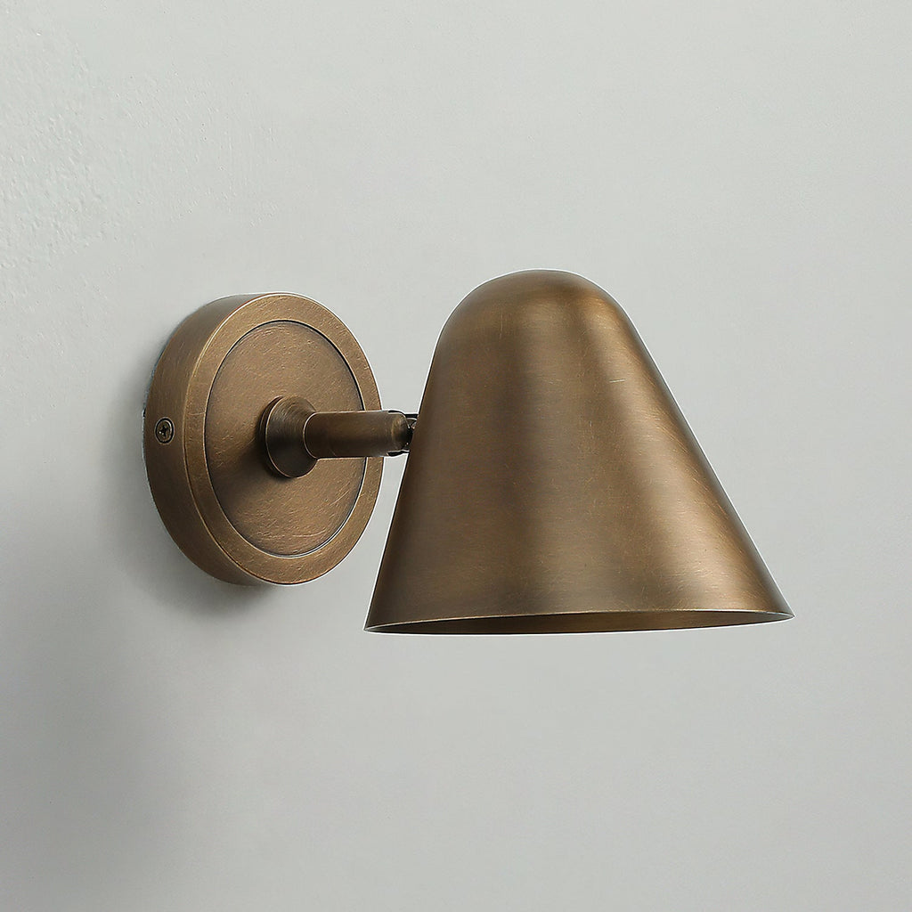 Side view of a Clérachic antique brass wall light with a round brass backplate, a short arm and a cone shade on a pale grey wall.