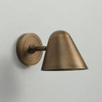 Side view of a Clérachic antique brass wall light with a round brass backplate, a short arm and a cone shade on a pale grey wall.