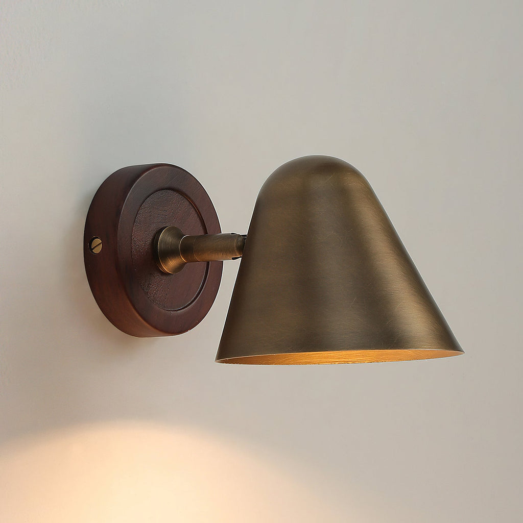 A Clérachic wall light with a cone shade in antique brass on a round walnut-coloured backplate, with warm light on the wall below.