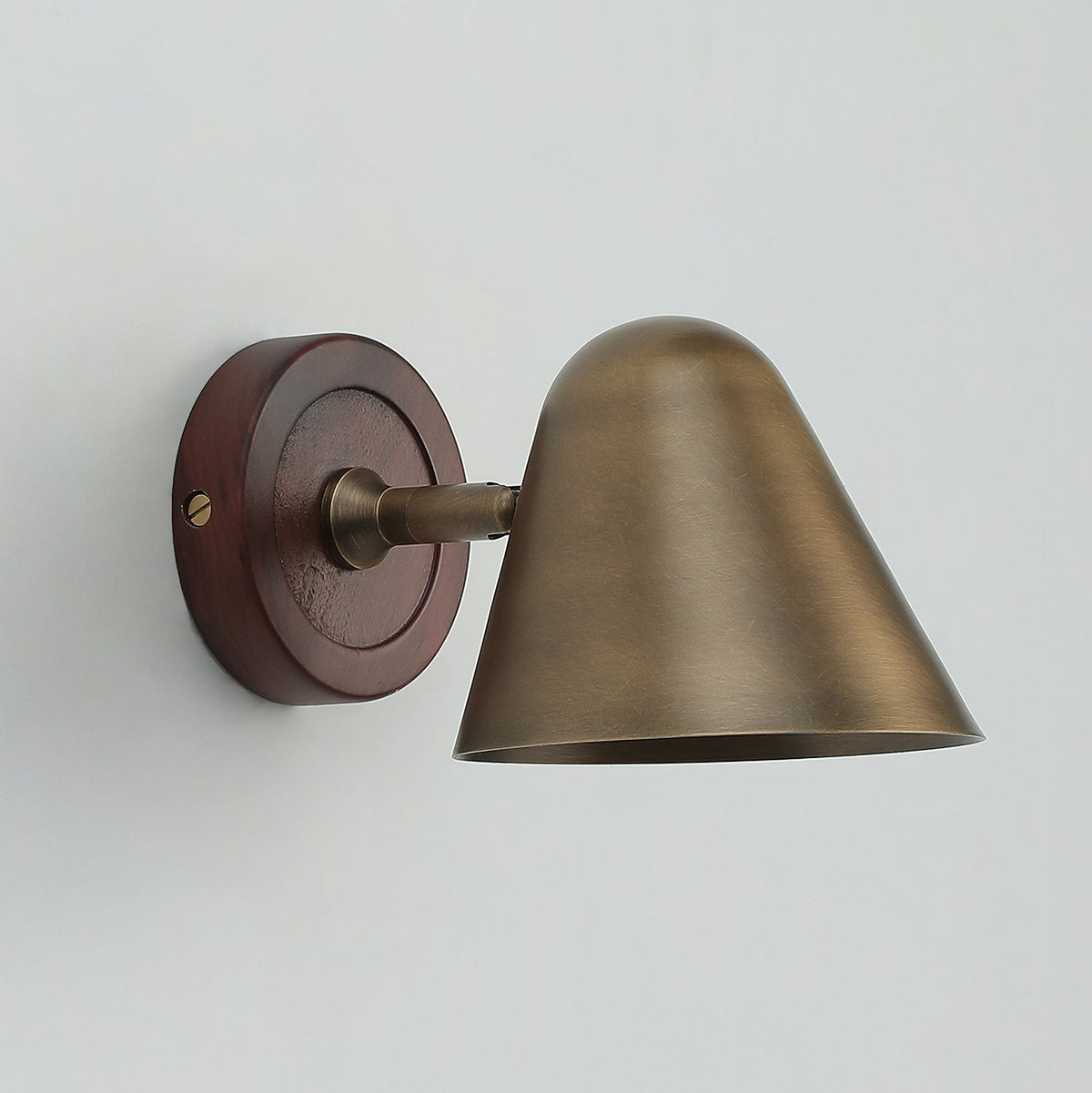 Side view of a Clérachic wall light with an antique brass cone shade and a round dark walnut-coloured backplate, on a pale wall.